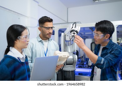 Caucasian Male Engineer Standing Hold A Robot Controller Next To Asian Female Apprentice Holding Laptop, Demonstrating The Work Of Robotic Machine To Young Asian Male CEO Who Looking Touching A Robot.