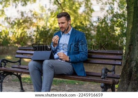 Caucasian male entrepreneur freelancer sitting outdoors in city park a bench talking online a video call using laptop or tablet. Business man office worker working on urban street background 