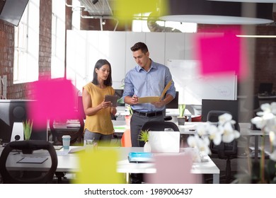 Caucasian Male And Female Colleague Discuss Tablet And File, View Through Glass Wall And Memo Notes. Working In Business At A Modern Office.