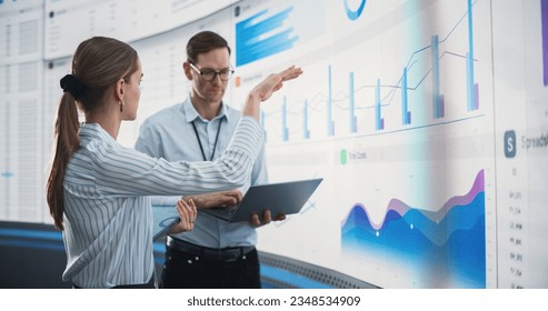 Caucasian Male And Female Data Scientists With Laptop And Tablet Standing Next To Big Digital Screen With Graphs And Charts In Monitoring Office. Colleagues Discussing Business Opportunities, Risks.