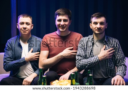 Caucasian male friends standing still in home when watching anthem at beginning sport game. Males football soccer fans singing hymn and holding hand on heart. Football fans watch match on TV