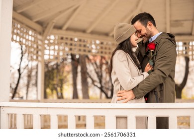 Caucasian male gifting a red rose to his girlfriend in a cozy pavilion. The couple enjoys a romantic moment, dressed in casual winter attire.  - Powered by Shutterstock