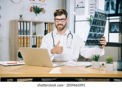 Caucasian Male Physician Smiling On Camera With Thumb Up, While Sitting At Desk With Tomography Scan In Hands. Bearded Doctor Wearing Eyeglasses, White Lab Coat And Using Laptop At Office.