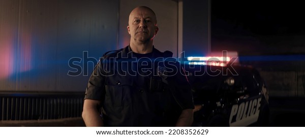 Caucasian male police\
officer posing against police car with flashing lights at night.\
Shot with anamorphic\
lens