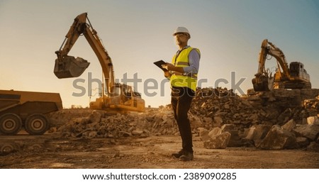 Caucasian Male Real Estate Investor Wearing Protective Goggles And Using Tablet On Construction Site On Sunny Day. Man Inspecting Building Progress. Excavator Loading Materials Into Industrial Truck