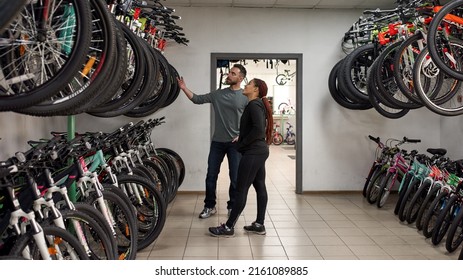 Caucasian male seller showing bicycles for smiling african american female customer in modern bike shop. Set of variety bikes for extreme sport, urban lifestyle and children. Shopping concept