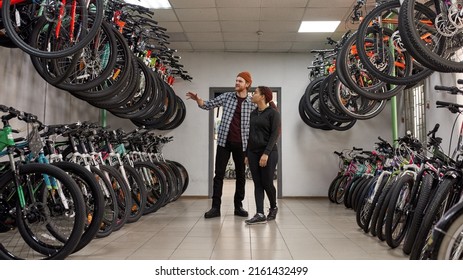Caucasian male seller showing bicycles for focused african american female customer in modern bike shop. Set of variety bikes for extreme sport, urban lifestyle and children. Shopping concept