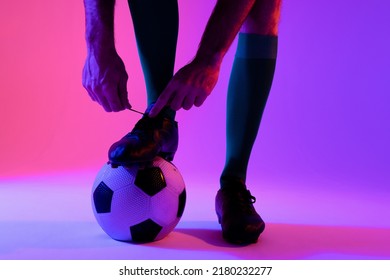 Caucasian Male Soccer Player With Football Tying Shoes Over Neon Pink Lighting. Sport, Movement, Training And Active Lifestyle Concept.