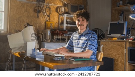 Caucasian Male Software Engineer Programming On Old Desktop Computer In Retro Garage, Looking At Camera And Smiling. Man Starting an Innovative Startup Company In Nineties. Nostalgia Concept.