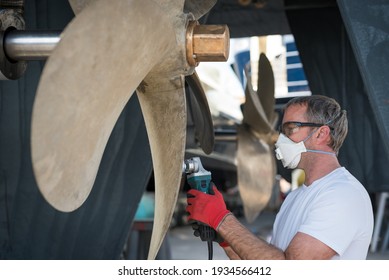 Caucasian male superyacht deckhand engineer polishing a yacht propeller with a power tool, gloves, mask and eye protection glasses 