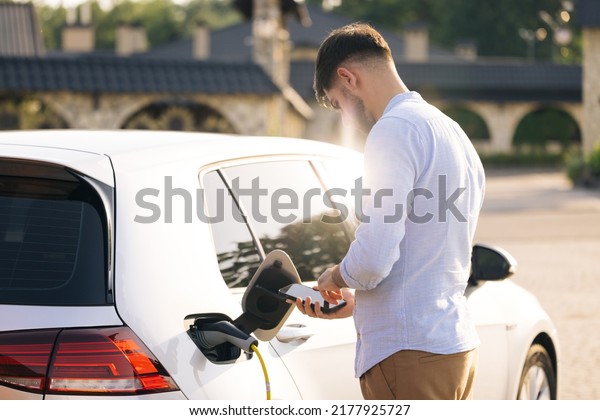 Caucasian male unplugging electric car from charging\
station. Man is unplugging in power cord to an electric car at\
sunset. Man charging electric car at charging station using smart\
phone app.