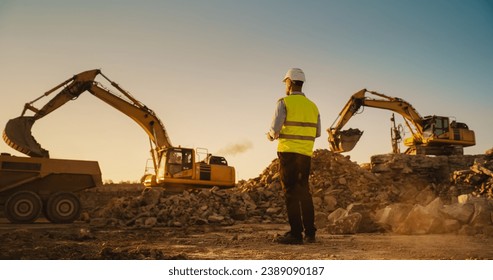 Caucasian Male Urban Planner Wearing Protective Goggles And Using Tablet On Construction Site On A Sunny Day. Man Inspecting Building Progress. Excavator Loading Materials Into Industrial Truck - Powered by Shutterstock