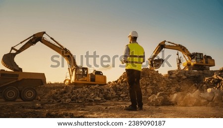 Caucasian Male Urban Planner Wearing Protective Goggles And Using Tablet On Construction Site On A Sunny Day. Man Inspecting Building Progress. Excavator Loading Materials Into Industrial Truck