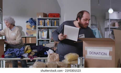 Caucasian Male Volunteer Writing On Clipboard While Working In Food Bank Warehouse. Young Fat Man Doing Inventory Of Donated Goods In Volunteer Center