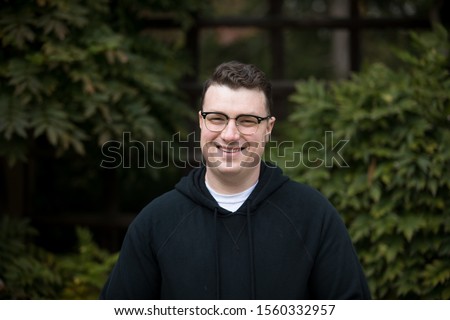 A caucasian male wearing a black hoodie sweater and glasses poses in a garden while surrounded by fall foliage on a cold afternoon.