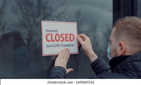 Caucasian Male Wearing Medical Mask Puts A Temporary Closed Due COVID-19 Pandemic Sign On A Window. Coronavirus Pandemic, Small Business Shutdown