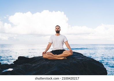 Caucasian Male Yogi Dressed In Comfortable Wear Meditating In Lotus Pose Enjoying Relaxation And Concentration Time At Seashore Rock, Concept Of Holistic Recovery And Mental Healthy Lifestyle