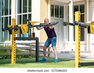 A Caucasian Man, 50 Years Old, Goes In For Sports, Trains Legs By Squatting With A Load, On An Open Sports Ground.