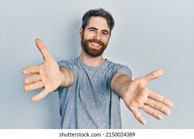 Caucasian man with beard wearing casual grey t shirt looking at the camera smiling with open arms for hug. cheerful expression embracing happiness. 