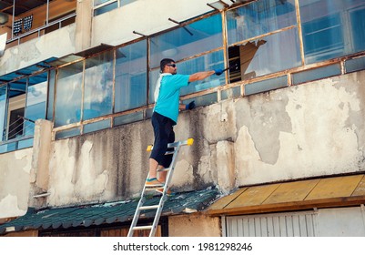 Caucasian Man Breaking A Window To Rescue His Neighbour Standing On A Ladder And Wearing A Protective Glove And Sunglasses