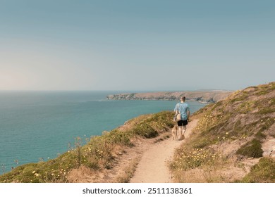 Caucasian man in casual attire walking Whippet dog along clifftop coastal path towards Bedruthan Steps with stunning sea views and green hills on a sunny day, overlooking the sea Cornwall, UK - Powered by Shutterstock