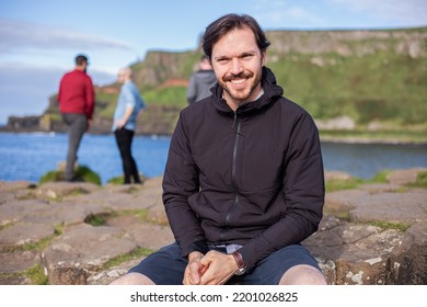 A Caucasian Man In Casual Black Clothes Sitting On Giant's Causeway Columns. Defocused People On Background. County Antrim, Northern Ireland