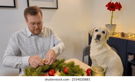 A Caucasian man crafting a Christmas wreath beside a Labrador, focusing on holiday preparation and festive decoration - Powered by Shutterstock