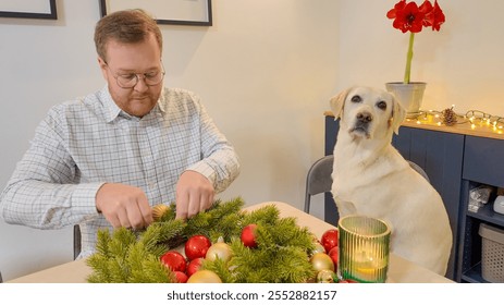 A Caucasian man crafts a Christmas wreath beside a curious Labrador, symbolizing holiday preparation and companionship - Powered by Shutterstock