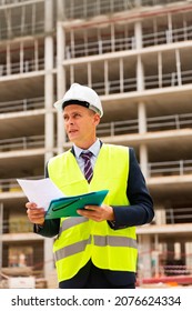 Caucasian Man Engineer In Workwear And White Hardhat Standing On Building Site And Analysing Documentation.