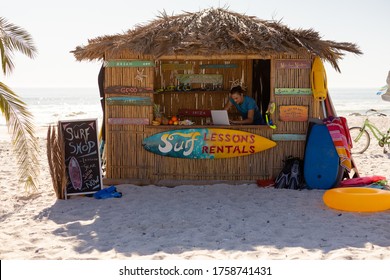 Caucasian man enjoying his time at the beach with his friends on a sunny day, using a laptop in a surfing lessons hut - Powered by Shutterstock