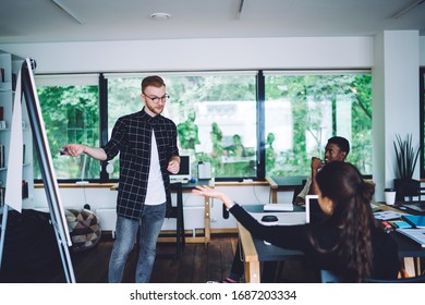 Caucasian Man Explaining Training Presentation To Freelance Colleagues During Collaboration In Modern Coworking Space With Flip Chart For Mentoring And Teaching, Group Of Smart Casual Millennials