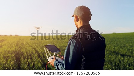 Caucasian man farmer in hat standing in green wheat field and controlling of drone which flying above margin. Male using tablet device as controller. Technologies in farming. Rear. Back view.