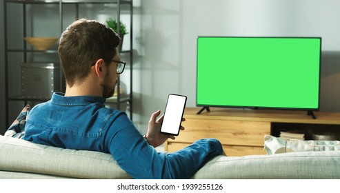 Caucasian man in glasses sitting on sofa in modern room in house watching TV with green screen and typing on smartphone with blank screen browsing online. Back view, leisure concept - Powered by Shutterstock