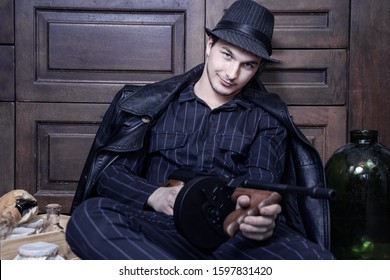 caucasian man with gun smiles sitting on the floor close to wine bottles and equipment. Gang or bootleggers wars concept during alcolol prohibition in USA in 1920-1930s. Classic striped suit and hat.