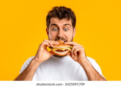 Caucasian man happily biting into a burger, portrait shot over yellow studio background, indulging in junk food, posing with delicious cheeseburger. Fast food enjoyment, nutrition habits - Powered by Shutterstock