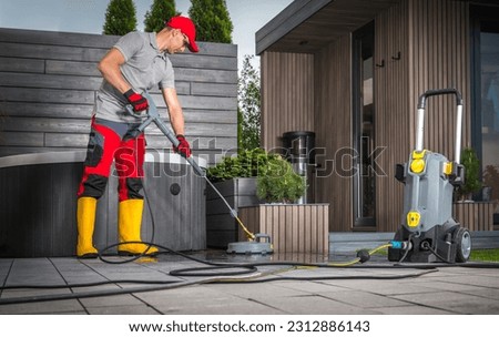 Caucasian Man in His 40s Pressure Washing Concrete Bricks Patio Using Surface Attachment. Modern Garden Shed and a Hot Tub in a Background.