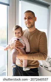 Caucasian Man Holding His Baby Girl Child Daughter By The Window At Home In Room In Day - Parenthood Care Father's Day Concept Real People Front View Looking To The Camera