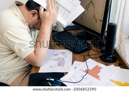 A Caucasian man holding his head worried about global economic losses with papers with printed calculation tables in his hands and on the desk. concept globalization, economy, frustration, anger.