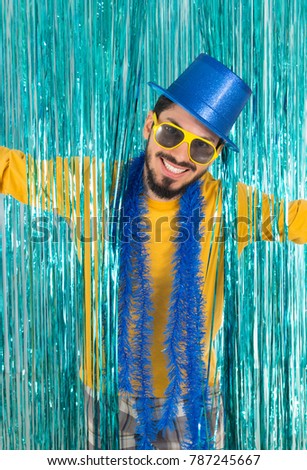 Caucasian man is holding open arms and smiling. Brazilian is wearing blue top hat, yellow sunglasses, and t-shirt. Bright curtain. Concept of new year eve, carnival and mardi gras.