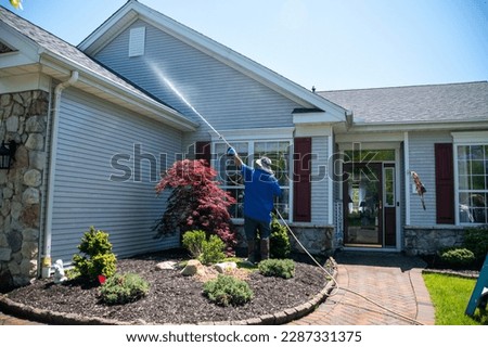 A caucasian man with a latex holding water sprayer wand power washing the siding of a house