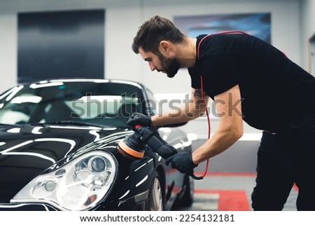 Caucasian man leaning over the front of black car using professional buffer tool to polish fresh paintwork. Car detailing process. Horizontal indoor shot. High quality photo