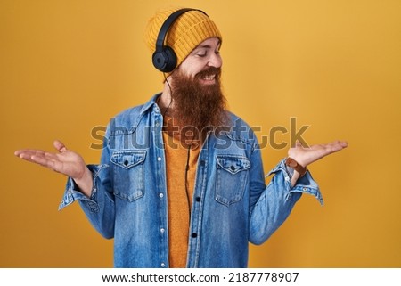 Caucasian man with long beard listening to music using headphones smiling showing both hands open palms, presenting and advertising comparison and balance 