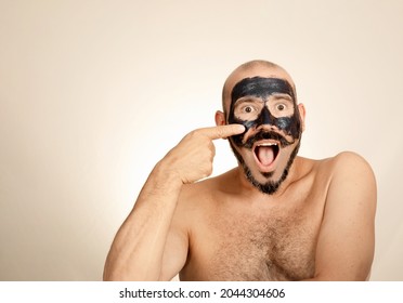 Caucasian Man Looks At Camera With Surprised Face While Applying Black Exfoliating Mask On His Face.  Bald Adult With Mustache Concept Health And Wellness, Skin Care.