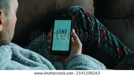 a caucasian man, lying in the sofa wearing pajamas and house robe, reads the text fomo, standing for fear of missing out, in his smartphone, in a panoramic format to use as web banner or header
