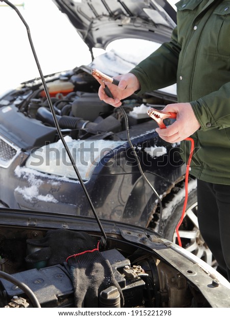 A Caucasian man mechanic stands with the starting\
wires near frozen snowy car engine, cold start with spare external\
battery accumulator and power wires with crocodile clips at Sunny\
winter day