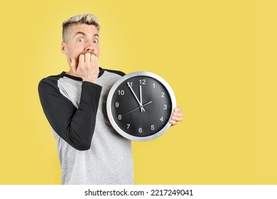 Caucasian Man On Casual Clothing, Eating Finger Nails While Holding Wall Clock, Worried For The Time. Studio Shot On Isolated Yellow Background. Copy Space On The Right Side