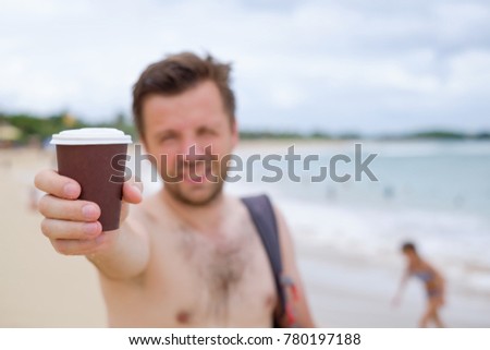 Caucasian man on summer beach suggesting cup of coffee. He is stretching his hand with mug ahead.