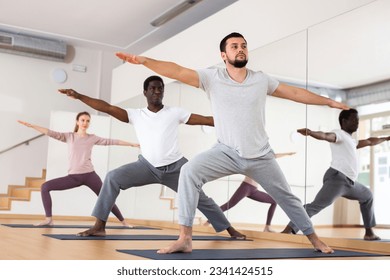 Caucasian man practising warrior II pose with people during group yoga training.