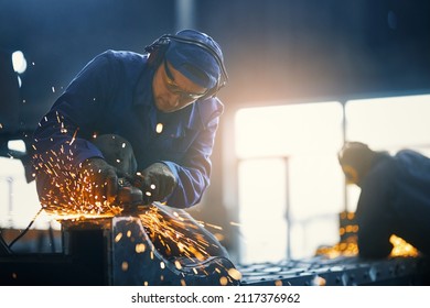 Caucasian man in protective clothes, glasses and gloves welding metal construction on factory. Professional locksmith making some details from steel at workshop. 