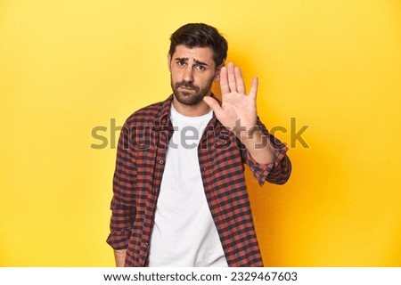 Caucasian man in red checkered shirt, yellow backdrop standing with outstretched hand showing stop sign, preventing you.